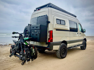 A beige Sprinter van parked on a sandy beach with a black roof rack, side windows, and a black utility box mounted on the rear. A black and green dirt bike is attached to the back of the van via an Atlas EBR1 rack